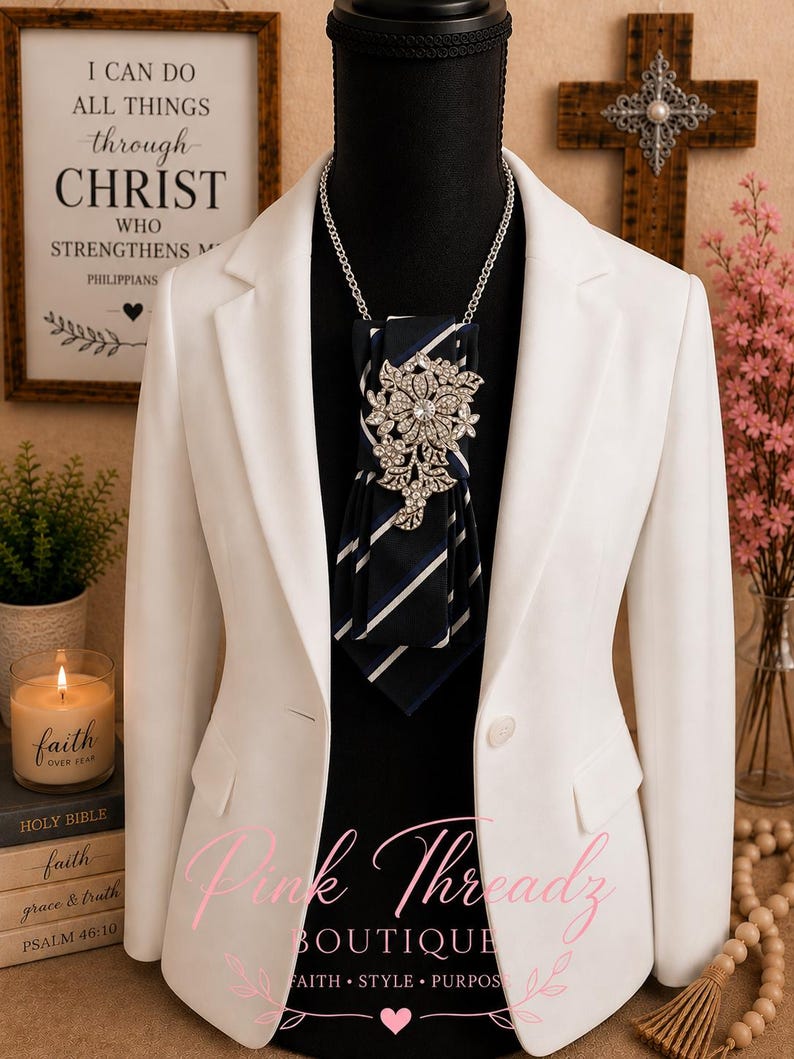 May include: A white blazer worn over a black mannequin, featuring a navy blue and white striped necktie accessory adorned with a large, ornate silver-toned floral brooch encrusted with clear crystals, suspended from a delicate silver-toned chain.