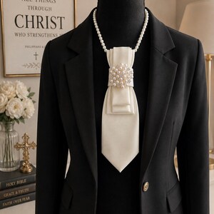 May include: A black blazer worn over a black garment, featuring a white satin necktie accessory. The tie is adorned with a central cluster of white pearls and clear crystals, suspended from a strand of white pearls worn around the neck.