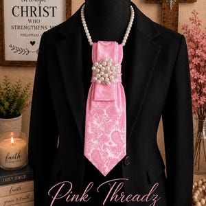 May include: A black blazer worn with a pink paisley patterned necktie featuring a large circular brooch made of white pearls and clear crystals. The tie is suspended from a white pearl necklace worn around the neck of a black mannequin torso.