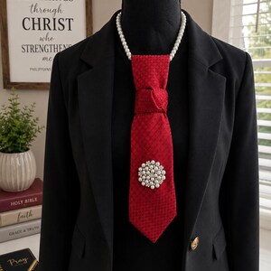 May include: A black blazer worn over a black top, featuring a red necktie with a subtle textured pattern. The tie is accented with a round brooch composed of small white pearls and clear crystals, and a strand of white pearls encircles the neck.