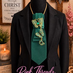May include: A dark green textured necktie featuring a gold chain and three cascading gold-tone butterfly brooches with green crystal accents. The tie is displayed on a black mannequin wearing a black blazer, with the text Pink Threadz BOUTIQUE.