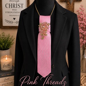 May include: A pink necktie with a subtle houndstooth pattern, featuring a gold-tone floral brooch with pink and clear crystals at the knot. The tie is displayed on a black mannequin wearing a black blazer, with the text Pink Threadz BOUTIQUE visible.