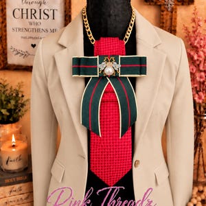 May include: A beige blazer worn over a red textured tie and a dark green bow tie featuring a central bee emblem with a white pearl. The bow tie has thin red stripes and gold trim. A gold chain necklace is visible around the neck area.