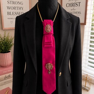 May include: A black blazer worn with a bright pink satin necktie featuring a gold chain necklace. The tie is adorned with two oval and circular cameo brooches in muted pink and gold tones, featuring intricate filigree details and small accents.