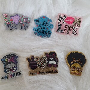 May include: Six colourful, glittery, resin-coated, decorative ornaments with various designs and text. The designs include a heart with the text "Love Mom Life", a peace sign, a heart with leopard print, a woman with a peace sign, a woman with a leopard print headband, and a zebra print with the text "Mom Life".