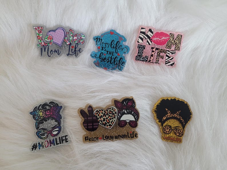 May include: Six colourful, glittery, resin-coated, decorative ornaments with various designs and text. The designs include a heart with the text "Love Mom Life", a peace sign, a heart with leopard print, a woman with a peace sign, a woman with a leopard print headband, and a zebra print with the text "Mom Life".