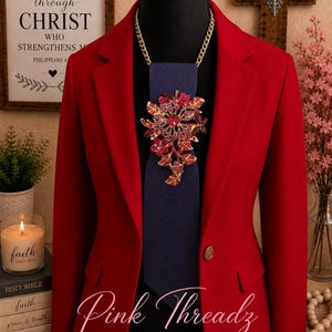 May include: A vibrant red blazer worn over a dark blue necktie. The tie is adorned with a large, ornate floral brooch featuring pink, purple, and red crystals. A gold-toned chain necklace secures the brooch and tie around the neck of the mannequin.
