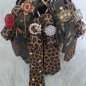 Peut inclure: Une collection de cravates à motifs léopard et camouflage, ornées de diverses broches décoratives incluant des cristaux rouges, des perles et des couronnes dorées. Les cravates sont superposées avec des chaînes dorées et des colliers de perles.