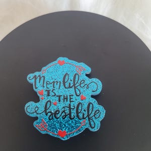May include: A blue glitter-covered sign with the text "Mom life is the best life" in black lettering. The sign is decorated with red hearts and arrows.