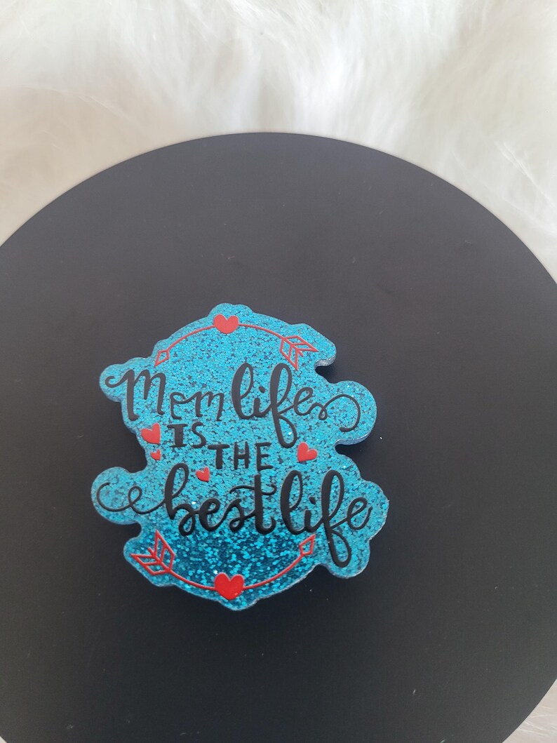 May include: A blue glitter-covered sign with the text "Mom life is the best life" in black lettering. The sign is decorated with red hearts and arrows.