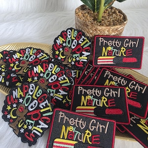 Puede incluir: Una colección de parches bordados con varios diseños. Algunos parches presentan el texto "NAPOLOGETICALLY" alrededor de un afro estilizado. Otros parches muestran el texto "Pretty Girl by Nature" con un gráfico de lápiz labial. Los parches son negros, rojos, amarillos y verdes.