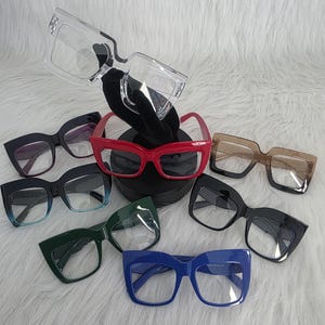 May include: Collection of square-framed eyeglasses in various colors, including clear, red, brown, black, green, and blue. The glasses have thick frames and are displayed on a white, textured surface.
