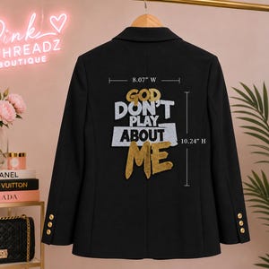 May include: A black blazer featuring a gold and white embroidered graphic on the back that reads GOD DON'T PLAY ABOUT ME. The design measures 8.07 inches wide by 10.24 inches high. The blazer has three gold buttons on each sleeve cuff.