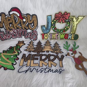 Puede incluir: Un conjunto de parches navideños con lentejuelas. Los diseños incluyen las palabras "Merry Christmas" con un gorro de Papá Noel, un árbol de Navidad, renos y la frase "Joy to the World". Los parches presentan lentejuelas doradas, rojas, verdes y azules.