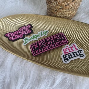 Pode incluir: Quatro patches bordados com texto em uma bandeja dourada em forma de folha. Os patches incluem frases como "They Not Like Us", "Beautiful Life", "I'm Not Perfect But I'm a Limited Edition" e "Girl Gang". Os patches são em várias cores.