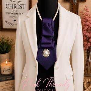 May include: A white blazer worn over a black mannequin, featuring a purple necktie with a textured fabric finish. The tie is adorned with a central oval brooch containing a white pearl surrounded by small clear crystals, paired with a pearl necklace.