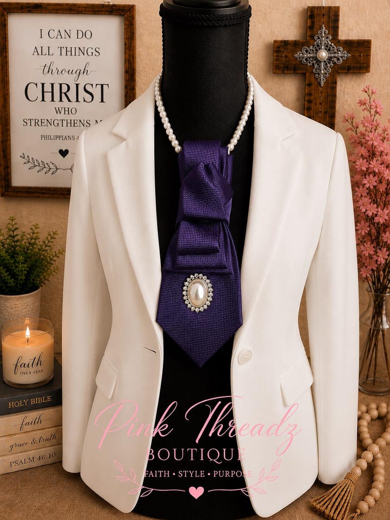May include: A white blazer worn over a black mannequin, featuring a purple necktie with a textured fabric finish. The tie is adorned with a central oval brooch containing a white pearl surrounded by small clear crystals, paired with a pearl necklace.