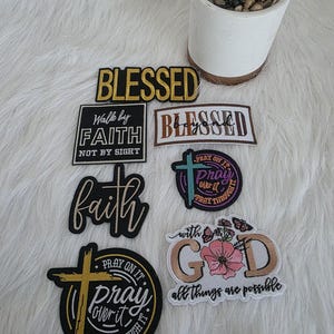 May include: A collection of embroidered patches with religious themes. The patches feature phrases like "Blessed," "Faith," and "Pray," along with floral and cross designs. The patches are in various colors, including black, gold, and white.