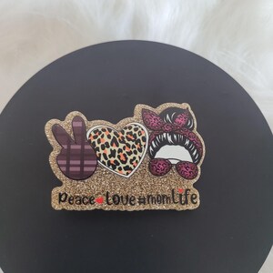 May include: Gold glitter peace sign, heart, and woman's face with sunglasses and a bow. The text "peace love #momlife" is below the design.