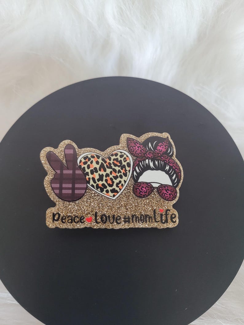 May include: Gold glitter peace sign, heart, and woman's face with sunglasses and a bow. The text "peace love #momlife" is below the design.