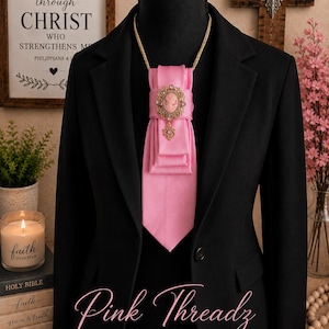 May include: A black blazer worn with a bright pink satin necktie featuring a gold-toned oval cameo brooch with intricate filigree detailing and a small dangling charm. The tie is secured with a gold chain around the neck of the display mannequin.
