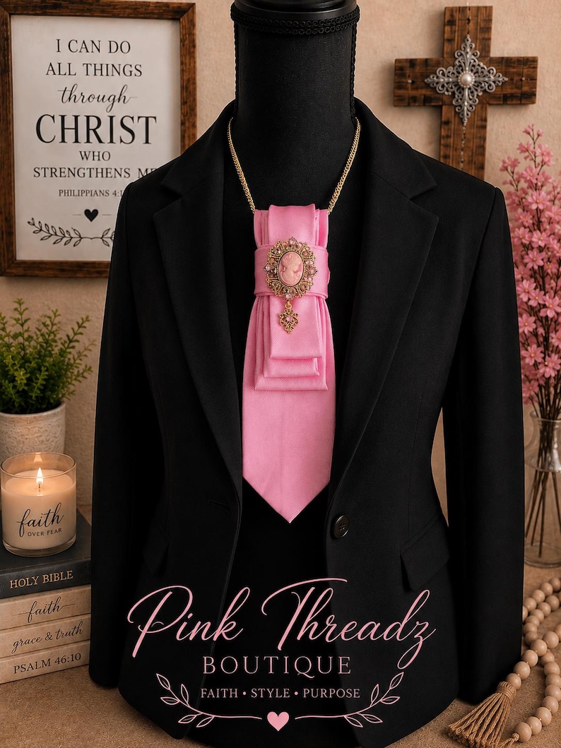 May include: A black blazer worn with a bright pink satin necktie featuring a gold-toned oval cameo brooch with intricate filigree detailing and a small dangling charm. The tie is secured with a gold chain around the neck of the display mannequin.