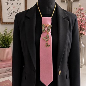 May include: A solid pink necktie featuring a gold-toned chain necklace and a vertical arrangement of three decorative butterfly brooches. The butterflies are embellished with green, pink, and gold-toned accents, centered on a black blazer.