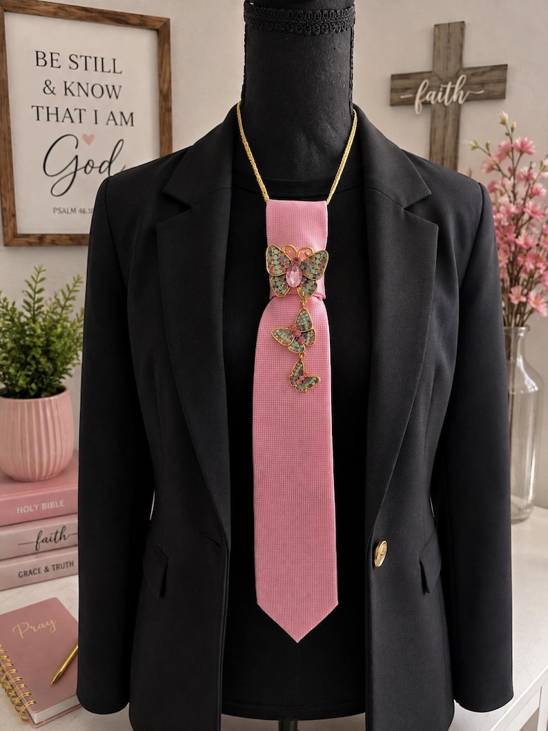 May include: A solid pink necktie featuring a gold-toned chain necklace and a vertical arrangement of three decorative butterfly brooches. The butterflies are embellished with green, pink, and gold-toned accents, centered on a black blazer.