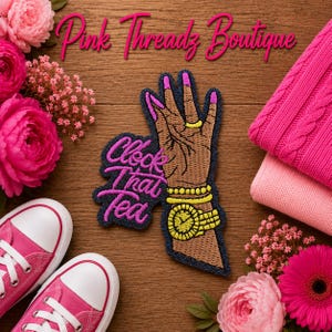 May include: An embroidered patch featuring a brown hand with pink manicured nails, wearing a yellow ring, yellow bracelets, and a yellow watch. The patch includes the text "Clock that Tea" in pink script. The background is a wood surface.