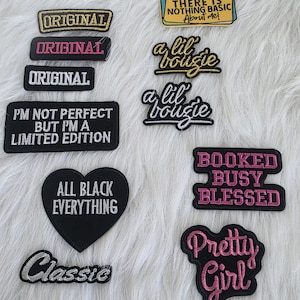 May include: A collection of embroidered patches in various shapes and colors. The patches feature phrases like "ORIGINAL," "a lil bougie," and "Pretty Girl." The patches are black, white, gold, and pink, with a heart-shaped patch that says "ALL BLACK EVERYTHING."