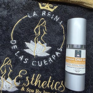 May include: A cylindrical bottle of Salmon DNA Serum with a silver cap and base, featuring a white label with black and orange text. The bottle is positioned on a black textured fabric embroidered with a gold crown, a lotus, and the text La Reina De Las Cuerpas Esthetics & Spa By Jamarie.