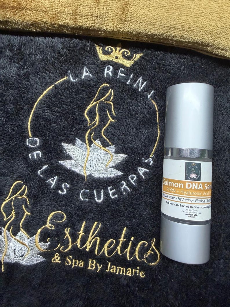 May include: A cylindrical bottle of Salmon DNA Serum with a silver cap and base, featuring a white label with black and orange text. The bottle is positioned on a black textured fabric embroidered with a gold crown, a lotus, and the text La Reina De Las Cuerpas Esthetics & Spa By Jamarie.