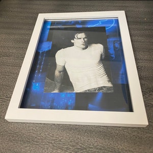 Puede incluir: Una foto en blanco y negro enmarcada de un hombre con una camiseta blanca y pantalones vaqueros. La foto está sobre un fondo azul con una imagen borrosa de una multitud en primer plano.