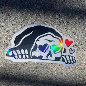 May include: A black and white sticker of a skull wearing a hooded cloak with rainbow hearts for eyes. The skull is looking to the right and has a small red heart and a smaller black heart floating above it.