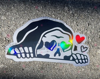 May include: A black and white sticker of a skull wearing a hooded cloak with rainbow hearts for eyes. The skull is looking to the right and has a small red heart and a smaller black heart floating above it.
