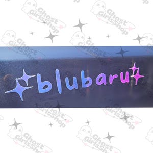 May include: A black surface with a pink and blue holographic sticker that says "#blubaru" with two sparkling stars on either side.