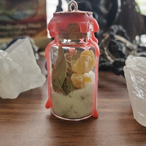 Pode incluir: Uma pequena garrafa de vidro com uma rolha de cortiça está cheia de sal branco, folhas verdes e resina amarela. A garrafa é selada com um selo de cera rosa.
