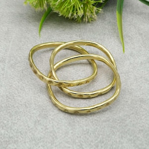 May include: Three gold-colored bangles with a textured, dotted pattern. The bangles are interlocked, creating a unique design. The jewelry is displayed on a light gray surface, with green foliage in the background.