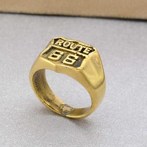 May include: A gold-colored ring with the words "ROUTE 66" and the number "66" embossed on the top. The ring has a classic signet style with a rectangular face and a smooth, polished band.