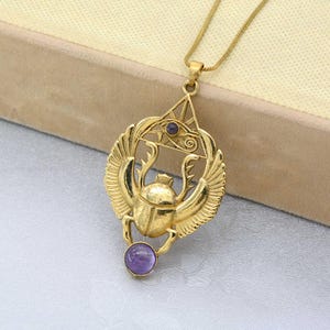 May include: Gold pendant necklace featuring a scarab beetle with wings, an eye of Horus, and a purple gemstone. The pendant is on a gold chain.