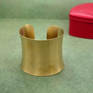 May include: A wide, gold-colored cuff bracelet with a brushed metal finish. The open-ended design features a curved shape, and the bracelet sits on a green surface. A red heart-shaped box is in the background.
