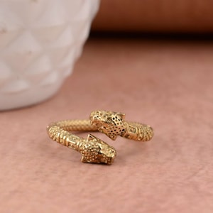 Op de afbeelding: Een goudkleurige ring met twee luipaardenkopontwerpen die tegenover elkaar staan. De ring heeft een getextureerde band en de luipaardenkoppen hebben gedetailleerde gelaatstrekken. De ring wordt getoond op een roze oppervlak.