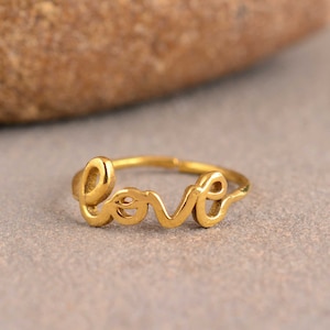 Puede incluir: Un anillo dorado con la palabra "love" escrita en letra cursiva.