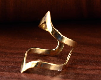 May include: A gold-colored ring with a unique, flowing design. The ring features a series of curved, wave-like shapes that create an open, artistic look. The metal has a polished finish, reflecting light and adding to its visual appeal. The ring is set against a dark wood surface.