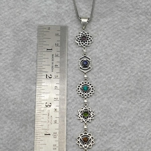 Op de afbeelding: Een zilverkleurige ketting met zeven chakrasymbolen, elk met een gekleurde edelsteen. De ketting is ongeveer 15 cm lang en is gemaakt in India.