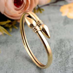 May include: A gold-coloured cuff bracelet with a distinctive design. The bracelet has two stylised, pointed ends that meet at the top. The metal has a polished finish, reflecting light. The bracelet is a piece of jewellery.