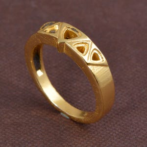 Puede incluir: Un anillo dorado con un diseño geométrico. El anillo presenta una serie de cortes triangulares a lo largo de la parte superior, creando un patrón calado. La banda es lisa y pulida, con una ligera textura visible en la superficie.