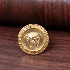 Peut inclure: Un bouton rond doré avec un motif de tête de Méduse en relief au centre. Le bouton est orné d'une bordure de clé grecque, sur un fond marron foncé.