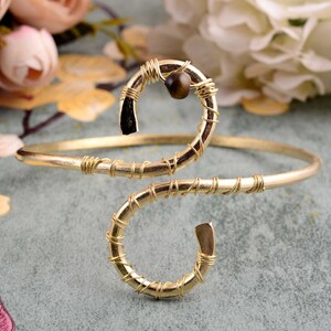 May include: A gold wire wrapped bracelet with a brown tiger's eye stone accent. The bracelet is made of two loops, one larger than the other, and is designed to fit snugly on the wrist.