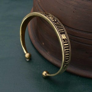 Könnte beinhalten: Ein goldfarbenes Metallarmband mit einem komplizierten geometrischen Muster. Das Armband ist offen und hat an beiden Enden eine abgerundete Form.